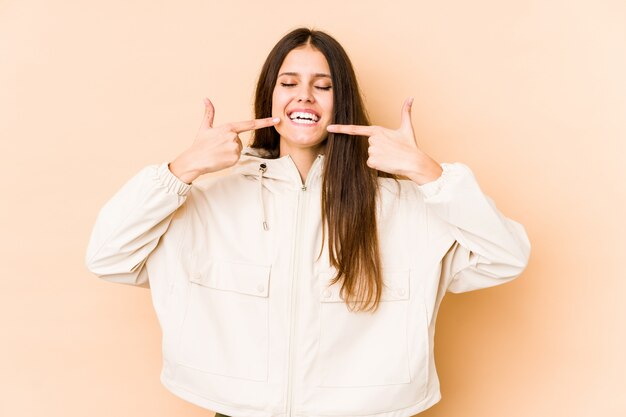 Giovane donna caucasica sui sorrisi beige della parete, indicando le dita alla bocca.