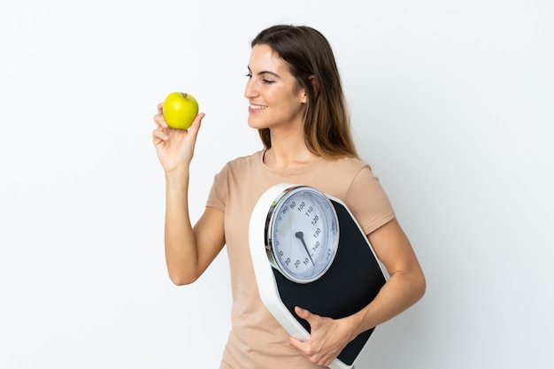 Giovane donna caucasica sul muro bianco con bilancia e con una mela