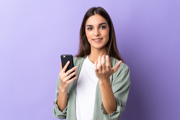 Giovane donna caucasica utilizzando il telefono cellulare isolato su sfondo viola che invita a venire con la mano. Felice che tu sia venuto