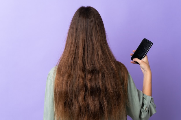 Giovane donna caucasica utilizzando il telefono cellulare isolato su sfondo viola nella posizione posteriore