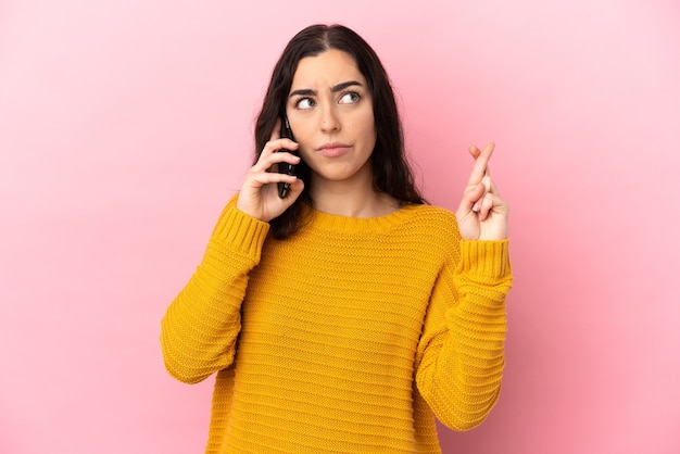 Giovane donna caucasica utilizzando il telefono cellulare isolato sulla parete rosa con le dita incrociate e augurando il meglio