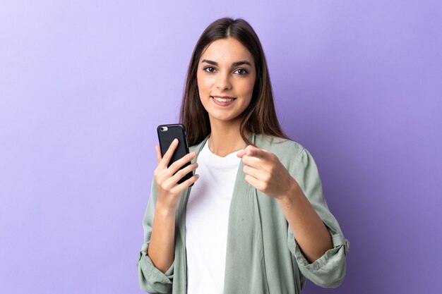 Giovane donna caucasica utilizzando il telefono cellulare su viola punta il dito contro di te con un'espressione fiduciosa