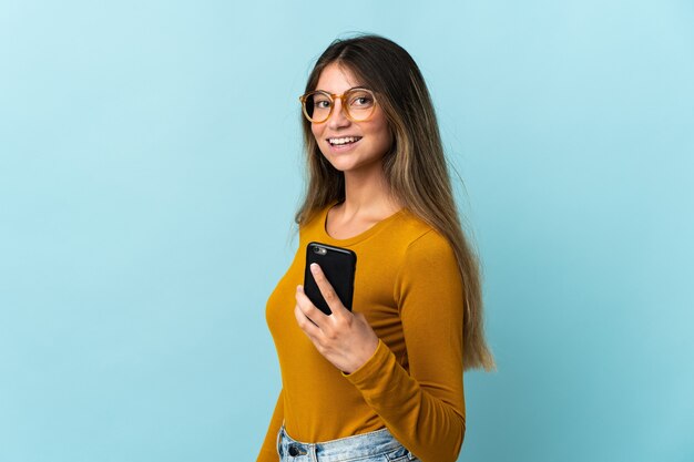 Giovane donna caucasica utilizzando il telefono cellulare sul blu che sorride molto