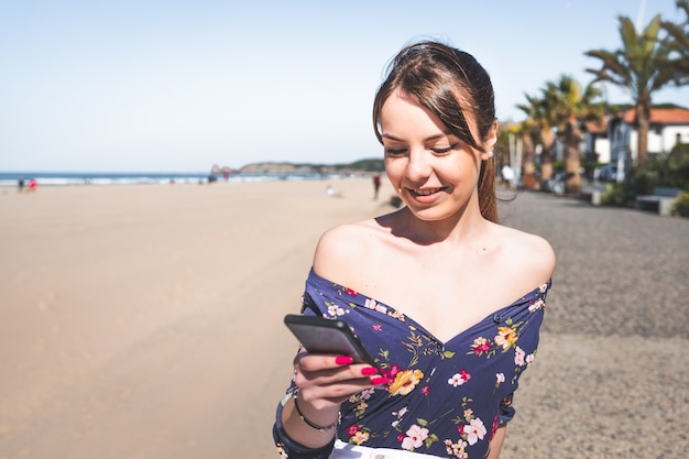Giovane donna caucasica utilizzando un telefono cellulare seduto sul lato di una passeggiata sulla spiaggia, a Hendaia, Paesi Baschi.