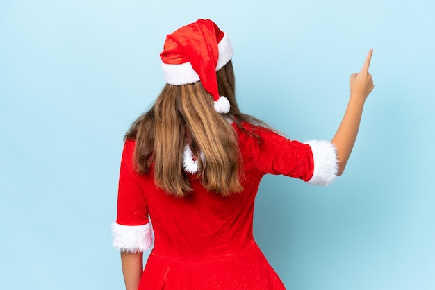 Giovane donna caucasica vestita da mamma noel isolata su sfondo blu che punta indietro con il dito indice