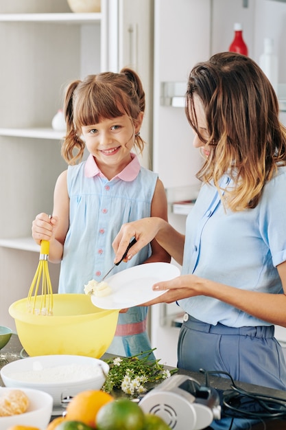 Giovane donna che aggiunge burro morbido in una grande ciotola di plastica quando sua figlia mescola gli ingredienti con la frusta