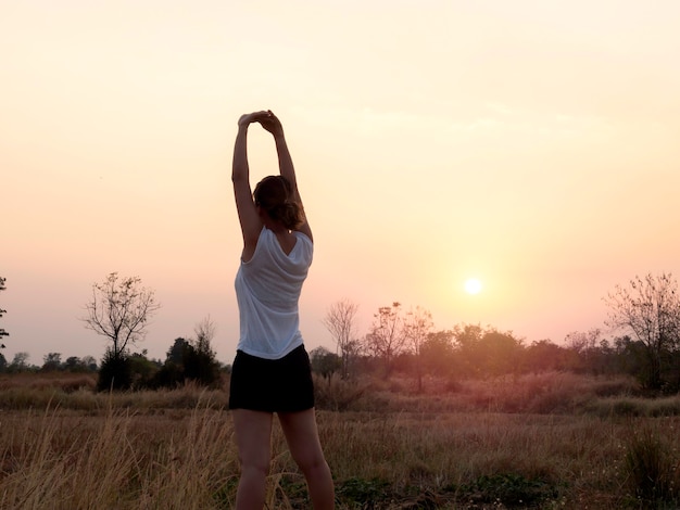 Giovane donna che allunga le braccia sul bellissimo cielo luminoso al tramonto vicino al campo estivo con spazio di copia