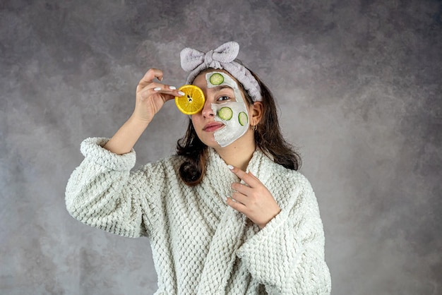 Giovane donna che applica una bella maschera di argilla bianca al viso con fette di cetriolo arancione isolato su grigio Procedura cosmetica di cura della pelle