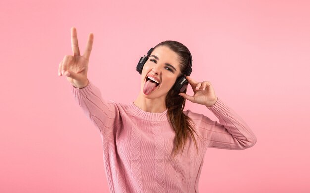 Giovane donna che ascolta la musica in cuffie senza fili che indossa un maglione rosa sorridente in posa sul rosa