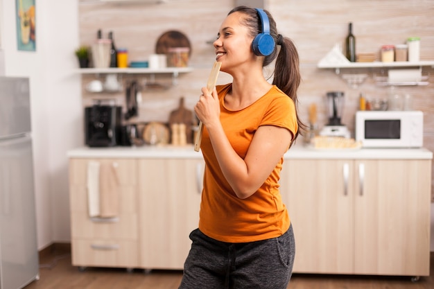 Giovane donna che ascolta musica sulle cuffie che canta sul cucchiaio di legno in cucina