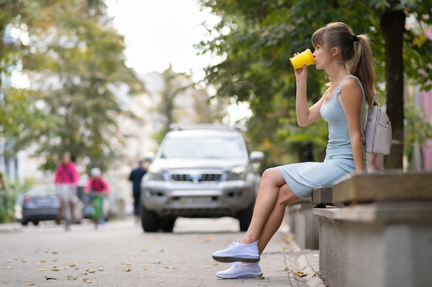 Giovane donna che beve caffè dal bicchiere di carta seduto sulla panchina della città nel parco estivo.