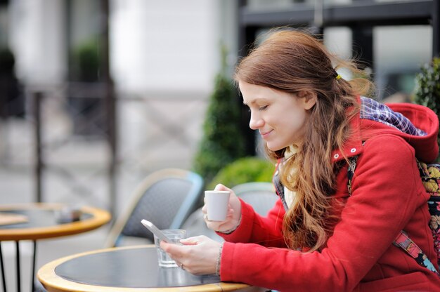 Giovane donna che beve caffè e usando il suo telefono intelligente in un caffè di strada parigino