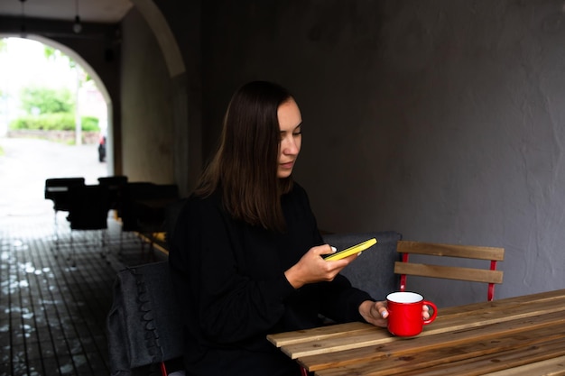 Giovane donna che beve da sola nella caffetteria sulla terrazza con uno smartphone in mano