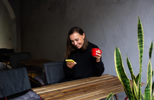 Giovane donna che beve da sola nella caffetteria sulla terrazza con uno smartphone in mano