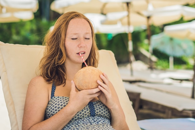 Giovane donna che beve latte di cocco sulla chaise-longue sulla spiaggia