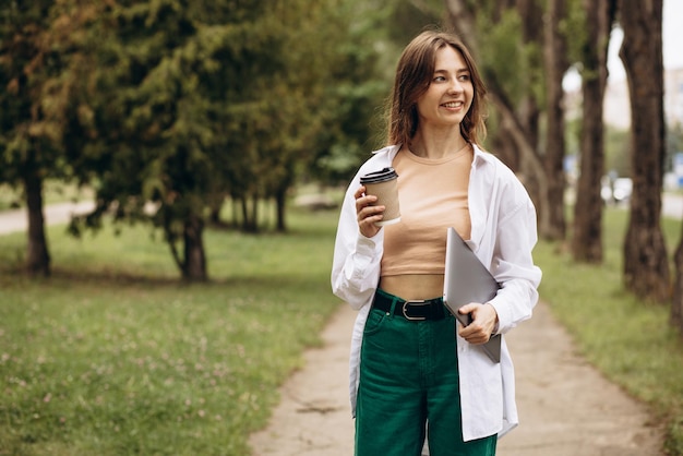 Giovane donna che cammina con il computer portatile nel parco e che beve caffè