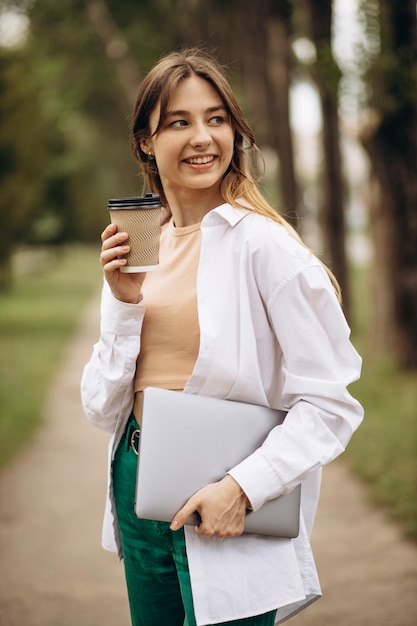 Giovane donna che cammina con il computer portatile nel parco e che beve caffè