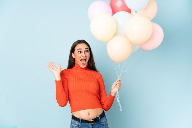 Giovane donna che cattura molti palloncini isolati sulla parete blu con espressione facciale a sorpresa