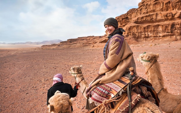 Giovane donna che cavalca un cammello nel deserto del Wadi Rum, guardando indietro sopra la sua spalla, sorridendo. Fa abbastanza freddo, quindi indossa il tradizionale cappotto beduino - bisht - e un foulard, sullo sfondo delle montagne