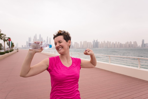 giovane donna che celebra un allenamento di successo eseguito sulla passeggiata in riva al mare con una bottiglia d'acqua e le mani alzate in aria con una grande città sullo sfondo