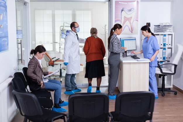 Giovane donna che chiede informazioni compilando un modulo stomatologico mentre i pazienti parlano seduti su una sedia nell'area di attesa. Persone che parlano nell'ufficio di reception dell'ortodontista professionale affollato.