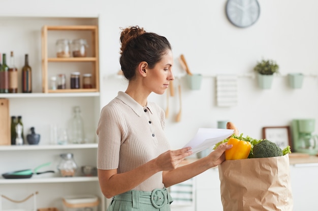 Giovane donna che controlla i prodotti dall'elenco dopo lo shopping in piedi in cucina