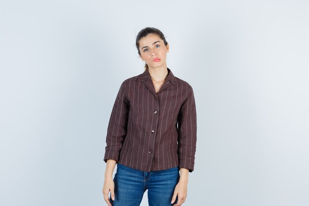 Giovane donna che curva le labbra mentre posa in camicia a righe, jeans e sembra carina. vista frontale.