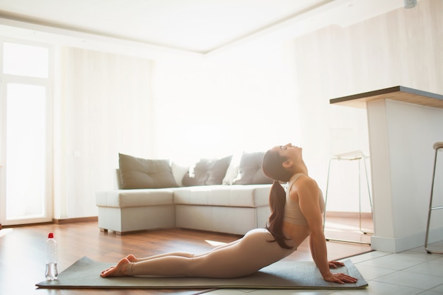 Giovane donna che fa allenamento di yoga nella sala durante la quarantena. Ragazza che fa posa rivolta verso l'alto del cane. Stretching schiena e parte superiore del corpo a casa sul tappetino.