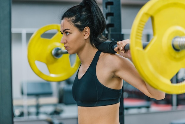 Giovane donna che fa allenamento in palestra spingendo il bilanciere giallo di sollevamento pesi sulle spalle Allenamento duro femminile per cross fit Persone sport fitness stile di vita sano e concetto di motivazione