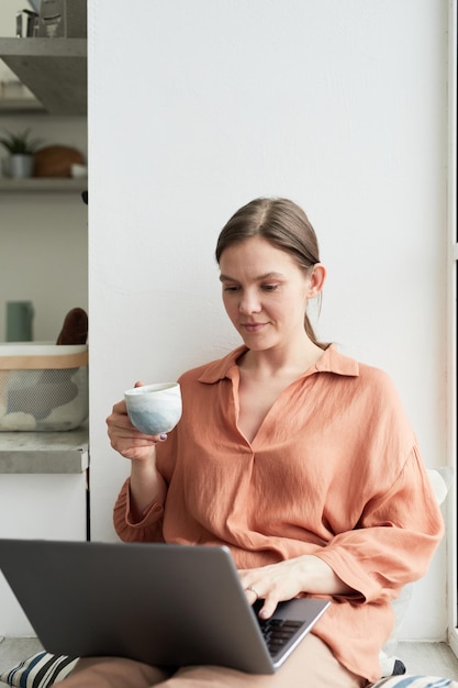 Giovane donna che fa il suo lavoro online sul laptop e beve caffè mentre è seduto nella stanza