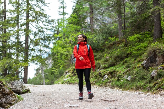 Giovane donna che fa un'escursione allegramente da sola nella foresta di montagna in un impermeabile rosso che guarda verso