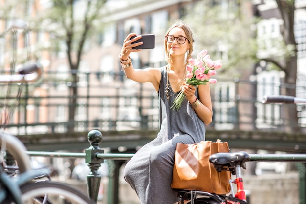 Giovane donna che fa una foto selfie con un mazzo di tulipani rosa seduto sul recinto vicino al canale d'acqua nella città di Amsterdam