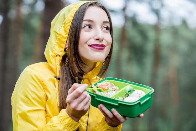 Giovane donna che fa uno spuntino con cibo sano nella scatola del pranzo durante il viaggio nella foresta