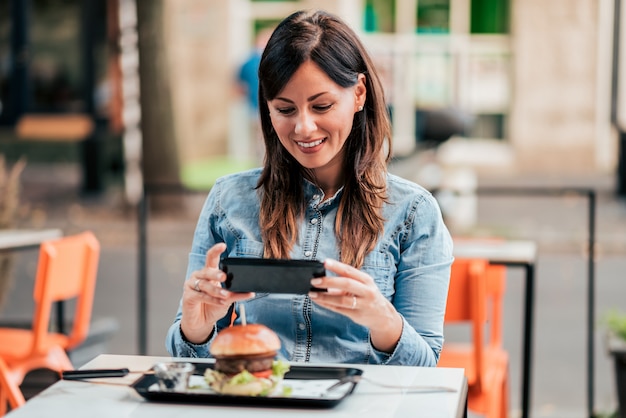 Giovane donna che fotografa hamburger delizioso nel ristorante di pasto rapido all&#39;aperto.