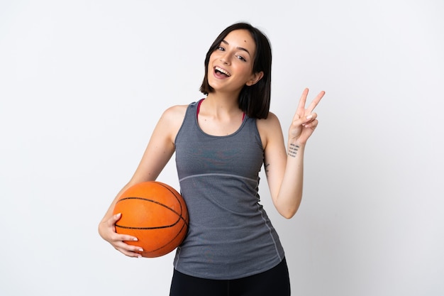 Giovane donna che gioca a basket isolato su sfondo bianco sorridendo e mostrando il segno di vittoria