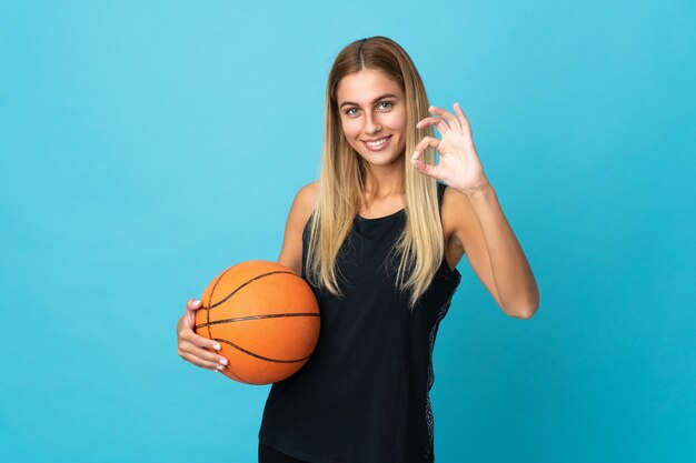 Giovane donna che gioca a basket isolato sul muro bianco che mostra segno ok con le dita