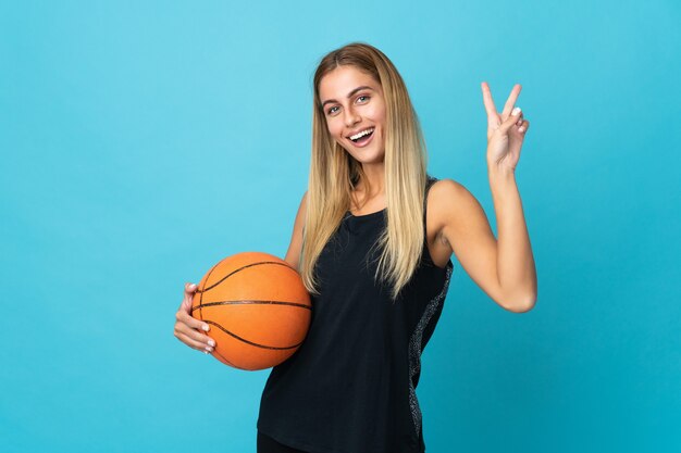 Giovane donna che gioca a basket isolato sul muro bianco sorridendo e mostrando il segno di vittoria