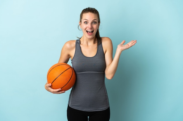 Giovane donna che gioca a basket isolato sull'azzurro con l'espressione facciale scioccata