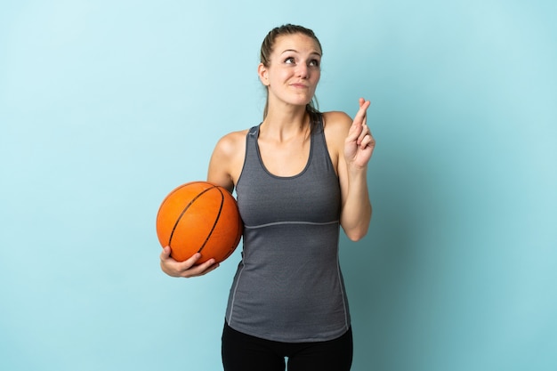 Giovane donna che gioca a basket isolato sulla parete blu con le dita incrociate e che desiderano il meglio