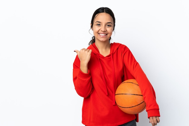 Giovane donna che gioca a basket sopra il muro bianco isolato che punta di lato per presentare un prodotto