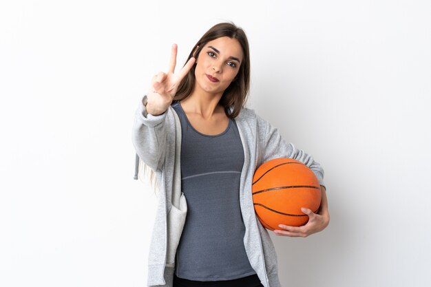 Giovane donna che gioca a basket su bianco sorridendo e mostrando il segno di vittoria