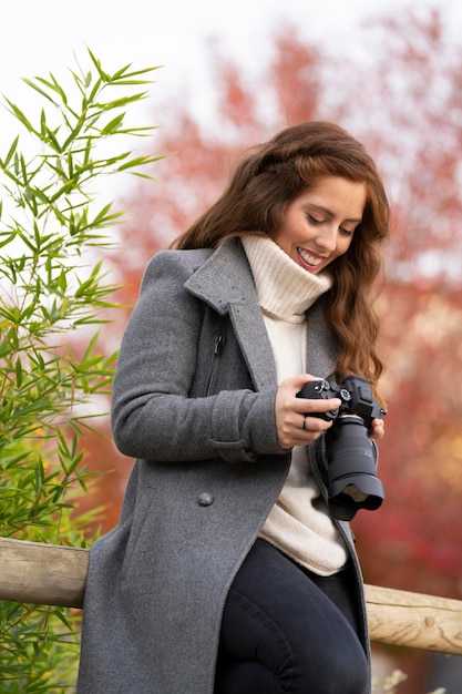 Giovane donna che guarda la sua macchina fotografica mirrorless e sorride in un parco Indossa un cappotto grigio e un maglione bianco Foto verticale