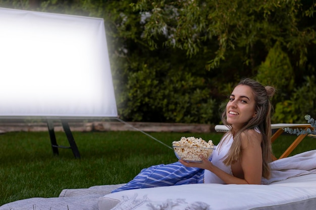 Giovane donna che guarda la telecamera godendosi il cinema estivo in giardino e tenendo una ciotola di popcorn