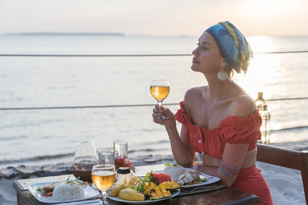 Giovane donna che ha una cena romantica nel ristorante dell'hotel durante il tramonto vicino alle onde del mare sulla spiaggia tropicale, primo piano. La ragazza si sta godendo il tramonto con un bicchiere di vino. Concetto di svago e viaggio