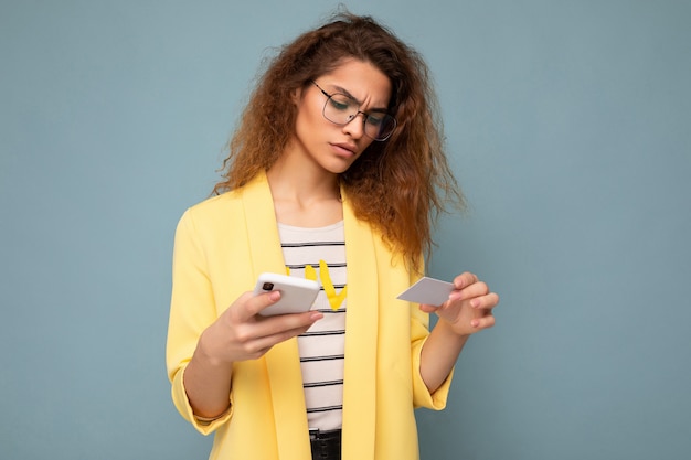 Giovane donna che indossa abiti di tutti i giorni isolati su sfondo tenendo il telefono e la carta di credito pagando lo shopping online tramite carta di credito guardando lo schermo dello smartphone