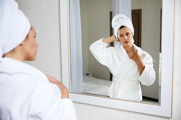 Giovane donna che indossa accappatoio con asciugamano sulla testa guardando lo specchio al mattino