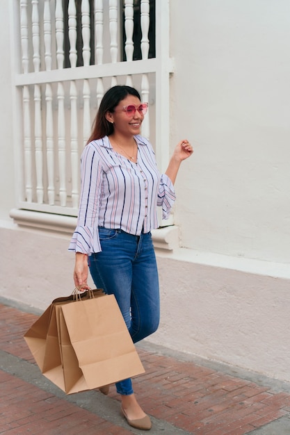 Giovane donna che indossa gli occhiali da sole che trasportano le borse della spesa mentre si sta in piedi vicino al muro