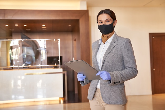 Giovane donna che indossa guanti di gomma e una maschera di tessuto mentre si trova in una hall di un hotel con un blocco per appunti