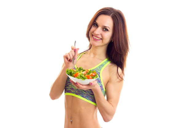 Giovane donna che indossa in cima sportiva colorata che tiene una forchetta e un piatto bianco con insalata verde e pomodoro