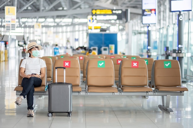 Giovane donna che indossa la maschera per il viso e seduta su una sedia in aeroporto, protezione dalla malattia da Coronavirus (Covid-19), viaggiatrice asiatica. New Normal, bolla di viaggio e allontanamento sociale